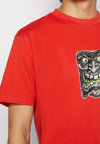 Camiseta de algodón roja con un gráfico de la cara de un monstruo con características exageradas y acentos verdes en la boca y los ojos.