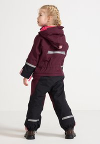 Bordeauxkleurige capuchonjack met roze voering, zwarte broek met reflecterende strepen en boorden. Buitenkleding voor actieve kinderen in de winter.
