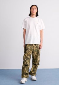 Young man standing wearing a plain white t-shirt, camouflage cargo pants, and white sneakers against a plain background.
