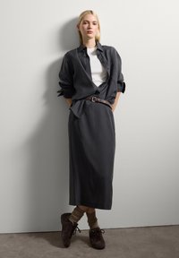 Jeune femme aux cheveux blonds s'appuie contre un mur blanc, portant une chemise noire à boutons, un haut blanc, une jupe noire, une ceinture marron, des chaussettes et des chaussures.
