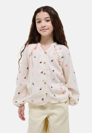 Jeune fille aux longs cheveux portant une blouse blanche à volants et une broderie florale colorée, associée à un pantalon clair.