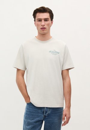 Lichtgrijs katoenen t-shirt met korte mouwen en een ronde hals. Heeft een blauwe tekstprint op de borst. Draagt samen met blauwe spijkerbroeken.