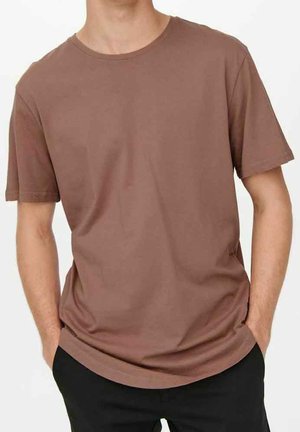 Homme portant un t-shirt marron uni à manches courtes, les mains dans les poches d'un pantalon noir, debout devant un fond blanc.