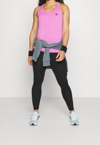 Top de deporte rosa con leggins negros, sudadera gris atada a la cintura, y zapatillas de deporte azul claro con detalles. Muñequeras negras en las muñecas.