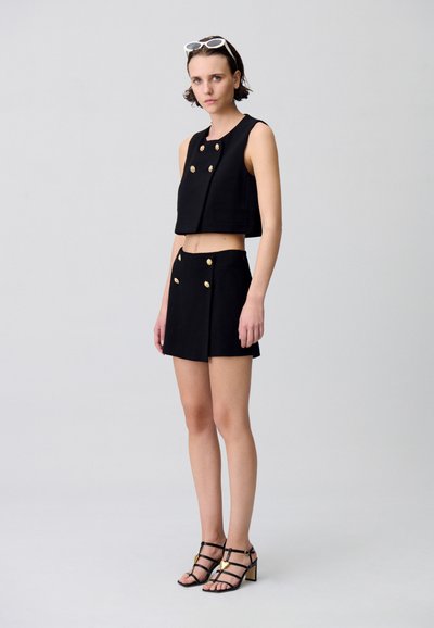 Haut noir court et short taille haute assorti avec des accents de boutons dorés. Le mannequin porte des talons noirs à lanières. Lunettes de soleil blanches comme accessoire.