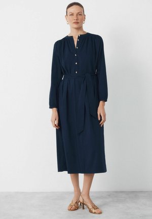 Femme portant une robe midi bleu marine à manches longues avec des boutons et une ceinture assortie, associée à des talons ouverts dorés, debout contre un mur blanc.