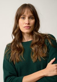 Maglione verde lavorato a maglia con scollo rotondo, texture morbida e vestibilità ampia; il modello ha lunghe onde di capelli castani e trucco neutro.