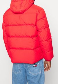 Rode pufferjas met horizontaal doorgestikt patroon en een capuchon. Kenmerkt zich door een soepele textuur en een elastische zoom. Draagt een paar blauwe denim jeans.