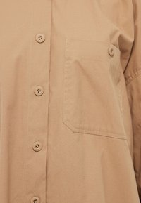 Camisa beige de botones con un bolsillo en el pecho, fabricada en un suave tejido de algodón, y cinco botones redondos con detalles cosidos. Textura suave en toda la prenda.