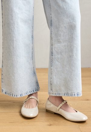 Sabrinas beijas com um salto baixo, apresentando um bico redondo e uma tira decorativa com pequenas pérolas. Usadas com jeans de perna larga azul claro.