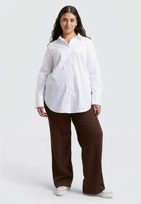 Chemise blanche boutonnée avec un col pointu, des manches longues et un ourlet arrondi, associée à un pantalon marron à jambes larges et des baskets blanches.
