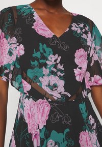 Robe noire à motifs floraux roses et teal. Comprend un décolleté en V, des manches transparentes et des accents en maille texturée.