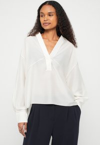 Hvit bluse med V-hals, plissert detalj langs halsen og lange, lett puffede ermer. Jevn tekstur, avslappet passform.