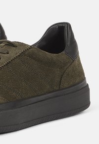 Olive green suede sneaker with a black leather heel, textured upper, flat laces, and a black rubber sole. Features diagonal stitching patterns.