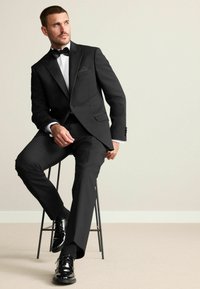 Tuxedo negru cu accente din satin, cămașă albă, papion negru. Model așezat pe un taburet metalic, pantofi negri lustruiți, fundal neutru.