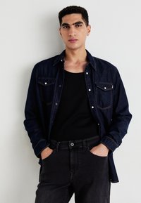 Tumman denim-nappipaidan edessä on nappikiinnitys ja kaksi rintataskua, yhdistettynä mustaan tank-toppiin ja tummiin farkkuihin. Korostaa kontrastivärin tikkausta.