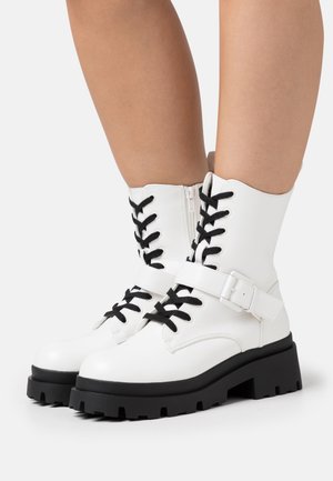 Valkoiset nilkkurit, joissa on edessä nyöritys, mustat nauhat, solkiremmi ja massiiviset mustat pohjat. Sileää synteettistä materiaalia, jossa mattapintainen viimeistely.