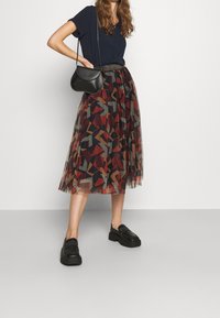 Chemise noire, jupe midi en tulle imprimée colorée avec des formes abstraites, ceinture élastique grise, et chaussures à plateforme noires. Détail du sac noir.