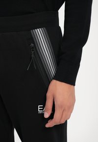 Close-up de uma mão próxima ao bolso de calças pretas com um fecho de correr vertical e um detalhe de faixa refletora ao longo da borda do bolso.