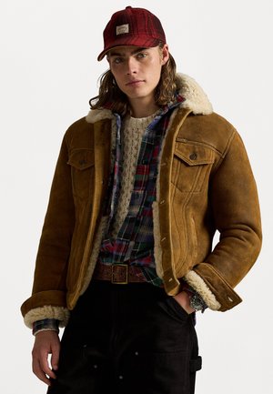 Chaqueta de ante marrón con collar de borreguito, superpuesta a una camisa de cuadros y un suéter de punto. Los accesorios incluyen una gorra de cuadros rojos y un reloj de pulsera.