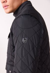 Black quilted jacket featuring a high collar, snap-button pockets, and an emblem on the shoulder. Smooth nylon texture, fitted design.