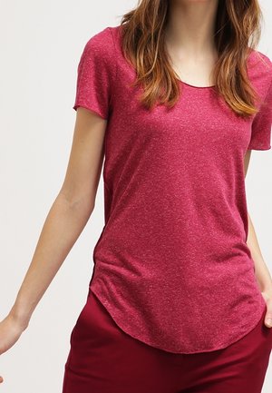 Kobieta ubrana w dopasowany, czerwony, melanżowy t-shirt z krótkim rękawem oraz pasujące czerwone spodnie, stojąca na tle jasnego, jednolitego tła.