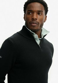 Czarny sweter dzianinowy z wysokim kołnierzem i półzamkiem, założony na zieloną i białą koszulę w paski. Posiada prążkowane detale przy kołnierzu.