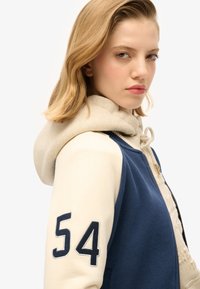 Sudadera con capucha de color azul en el cuerpo y mangas crema. Presenta un cierre vertical, un gran número "54" en la manga y una textura de tela suave.