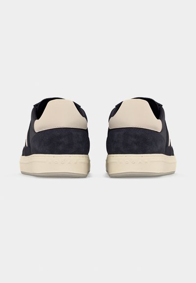 Ζευγάρι μαύρα και μπεζ αθλητικά παπούτσια με λευκές σόλες, εμφανιζόμενα από πίσω σε απλό φωτεινό φόντο.