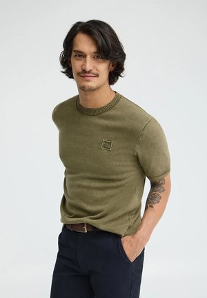 Uomo con capelli mossi scuri che indossa un maglione verde oliva a maniche corte e pantaloni scuri, con una mano in tasca su uno sfondo uniforme.