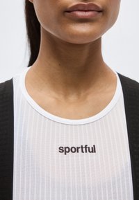 Κοντινό πλάνο ενός ατόμου που φοράει λευκή αθλητική μπλούζα με μαύρες τιράντες, με τη λέξη "sportful" τυπωμένη στο στήθος.