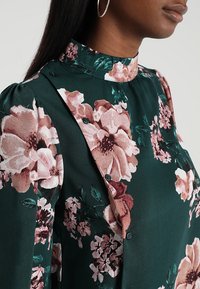 Blusa con stampa floreale su sfondo verde scuro, con grandi fiori rosa e crema, colletto alto e dettagli con bottoni sul davanti.