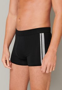 Černé boxerky vyrobené z pružného materiálu, s měkkým pasem a dvěma šedými vertikálními pruhy po stranách.