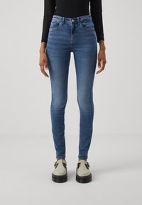 Középkék farmer skinny farmernadrág, közepes derékvonallal, öt zsebes kialakítással és enyhe kopással. Szürke talpkiemelő cipővel párosítva.