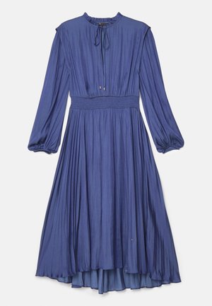 Kongelig blå, plisseret kjole med høj hals og lange ærmer, parret med hvide slingback hæle. Den glatte stof kontrasterer med den teksturerede stol.