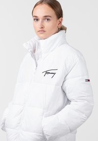 Bijela puffer jakna s patentnim zatvaračem sprijeda, visokim ovratnikom i izvezenim "Tommy" logotipom. Ima trobojne akcentne detalje na rukavu i kviltanu teksturu.