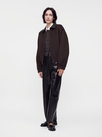 Άτομο που στέκεται φορώντας ένα σκούρο καφέ oversized σακάκι, μαύρο πουκάμισο με κουμπιά, μαύρο δερμάτινο παντελόνι και μαύρα μοκασίνια, μπροστά σε απλό λευκό φόντο.