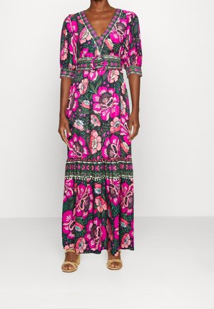 Femme portant une longue robe fleurie rose et verte à manches mi-longues avec une taille ceinturée assortie, assortie à des sandales ouvertes beige.