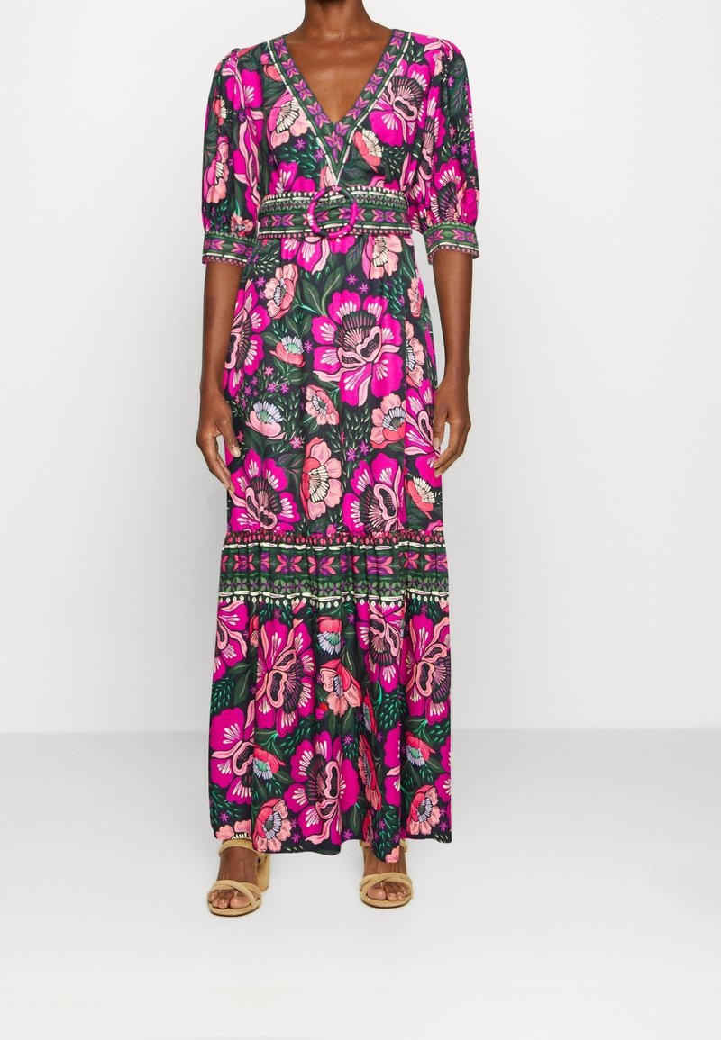 Femme portant une longue robe fleurie rose et verte à manches mi-longues avec une taille ceinturée assortie, assortie à des sandales ouvertes beige.