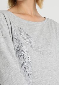 Sudadera de algodón gris con un diseño texturizado y acentos de plumas en lentejuelas plateadas en el frente. Tela suave, escote amplio, corte relajado.