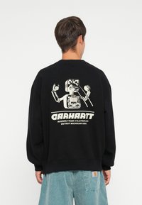 Zwarte sweater met ronde hals, voorzien van een grafisch ontwerp van een robot op de achterkant, vergezeld van de tekst "Carhartt" en details.