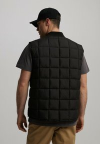 Czarna kamizelka puchowa w kratkę, wykonana z błyszczącego materiału izolacyjnego. Nosi się ją na koszulce z krótkim rękawem w kolorze szarym, która nie ma rękawów.