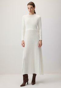 Vrouw met bruin haar in een paardestaart, gekleed in een lange witte ribbelgebreide jurk met een geplooide rok en bruine hoge hakken, staande voor een effen achtergrond.