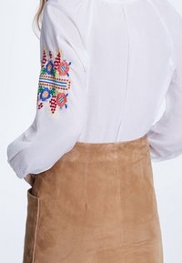 Blouse blanche avec des motifs floraux brodés colorés sur la manche gauche, associée à une jupe en daim marron clair avec une poche latérale.