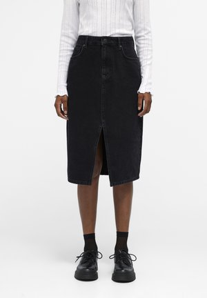 Black denim skirt with a front slit, high waist, and belt loops. Paired with a long-sleeve white shirt and black shoes.