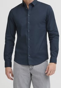 Overhemd met lange mouwen en knopen in een donker marineblauwe stof. Bevat een klassieke kraag, rechte zoom en knopen aan de voorkant. Draagt met grijze jeans.