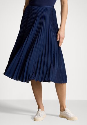 Jupe plissée bleu marine avec une texture lisse et une ceinture, associée à des baskets de couleur claire. La jupe arrive au milieu du mollet.