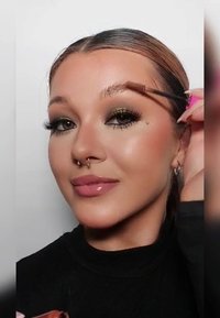 Personne appliquant un gel à sourcils avec un pinceau. Présente un teint clair, un eye-liner subtil et des lèvres brillantes. Porte un haut noir et des boucles d'oreilles créoles.