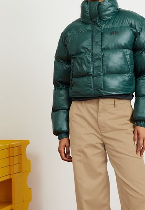 Persoon die een donkergroen donsjack met het "HH"-logo en beige broek draagt, staand bij een gele geribbelde opslagunit tegen een lichte achtergrond.