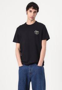 Black cotton t-shirt with short sleeves, featuring a small printed logo in white and green on the left chest, paired with blue denim pants.